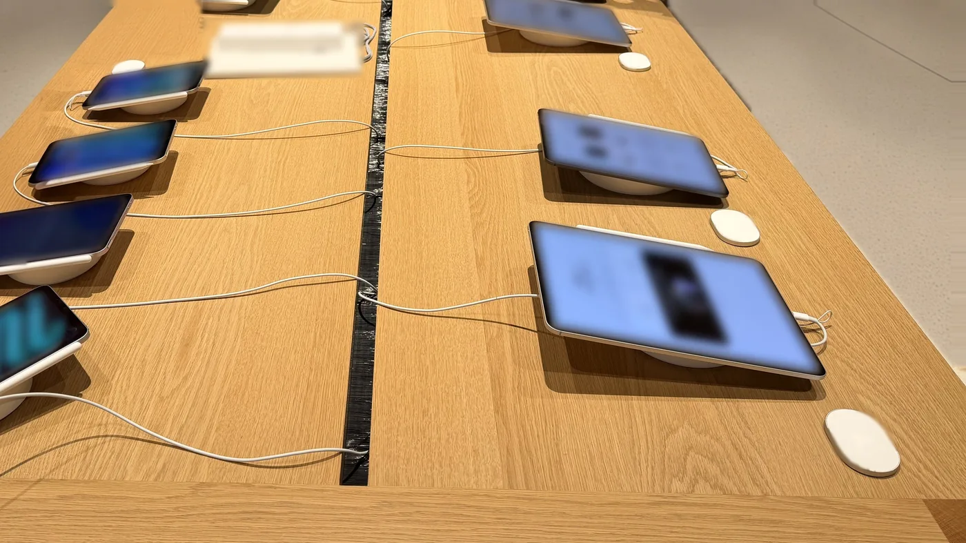 Style reference photo taken at an Apple Store: wooden display table with iPads on round stands, each tethered by a slim charging cable (not Tocvue products)